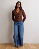 A woman stands against a textured off-white wall on a light wood floor, wearing a brown long-sleeve button-front top with gathered ruffle details down the front and wide-leg blue jeans.