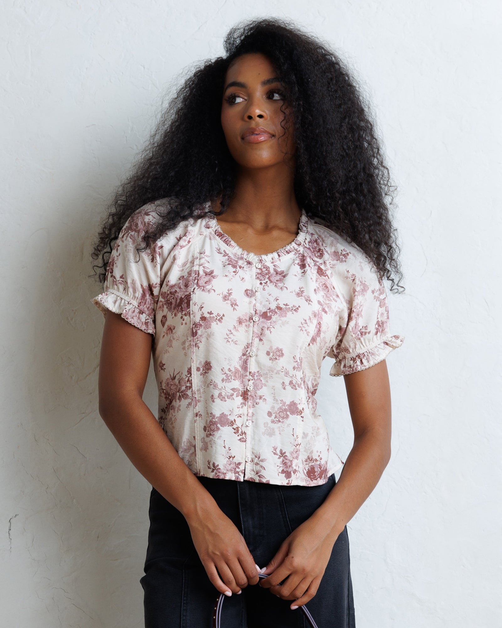 A woman with long, curly black hair wearing a short-sleeve blouse with a dusty rose floral print on a cream background, paired with dark jeans, standing against a textured white wall.