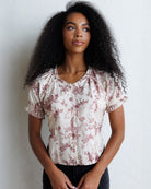 A woman with curly black hair wearing a short-sleeve cream blouse with a dusty rose floral print and puff sleeves, standing against a white textured wall.