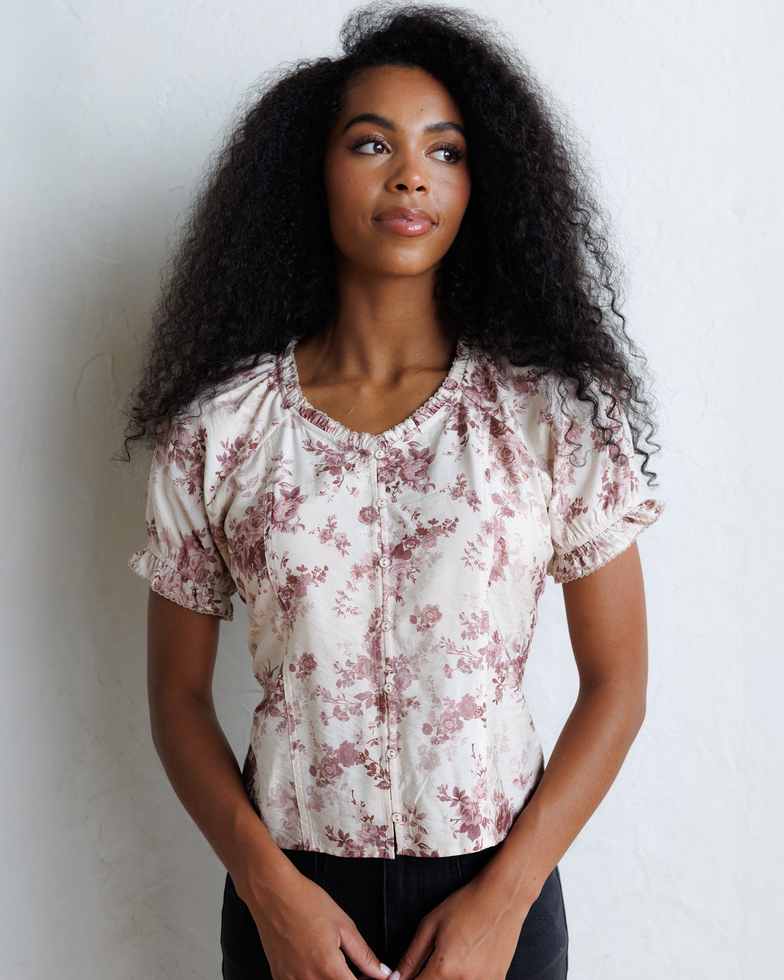 A woman with curly black hair wearing a short-sleeve cream blouse with a dusty rose floral print and puff sleeves, standing against a white textured wall.