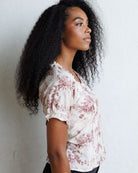 A woman with long, curly black hair is shown in profile wearing a short-sleeve blouse with a dusty rose floral print on a cream background, standing against a light textured wall.