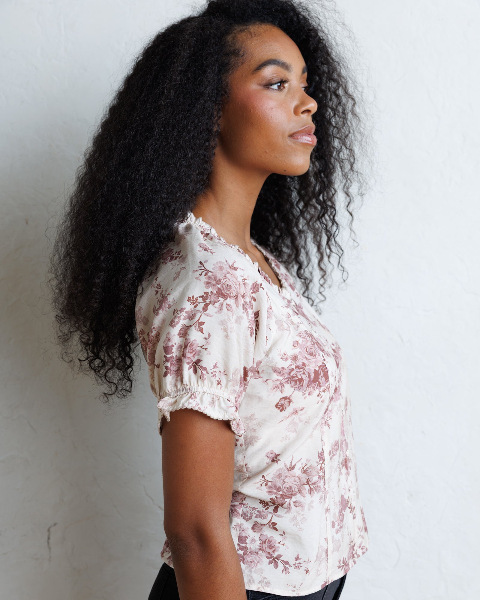 A woman with long, curly black hair is shown in profile wearing a short-sleeve blouse with a dusty rose floral print on a cream background, standing against a light textured wall.