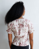 A woman viewed from behind wearing a cream-colored blouse with a dusty rose floral pattern, short puff sleeves, and a smocked waist, paired with black pants against a white wall.