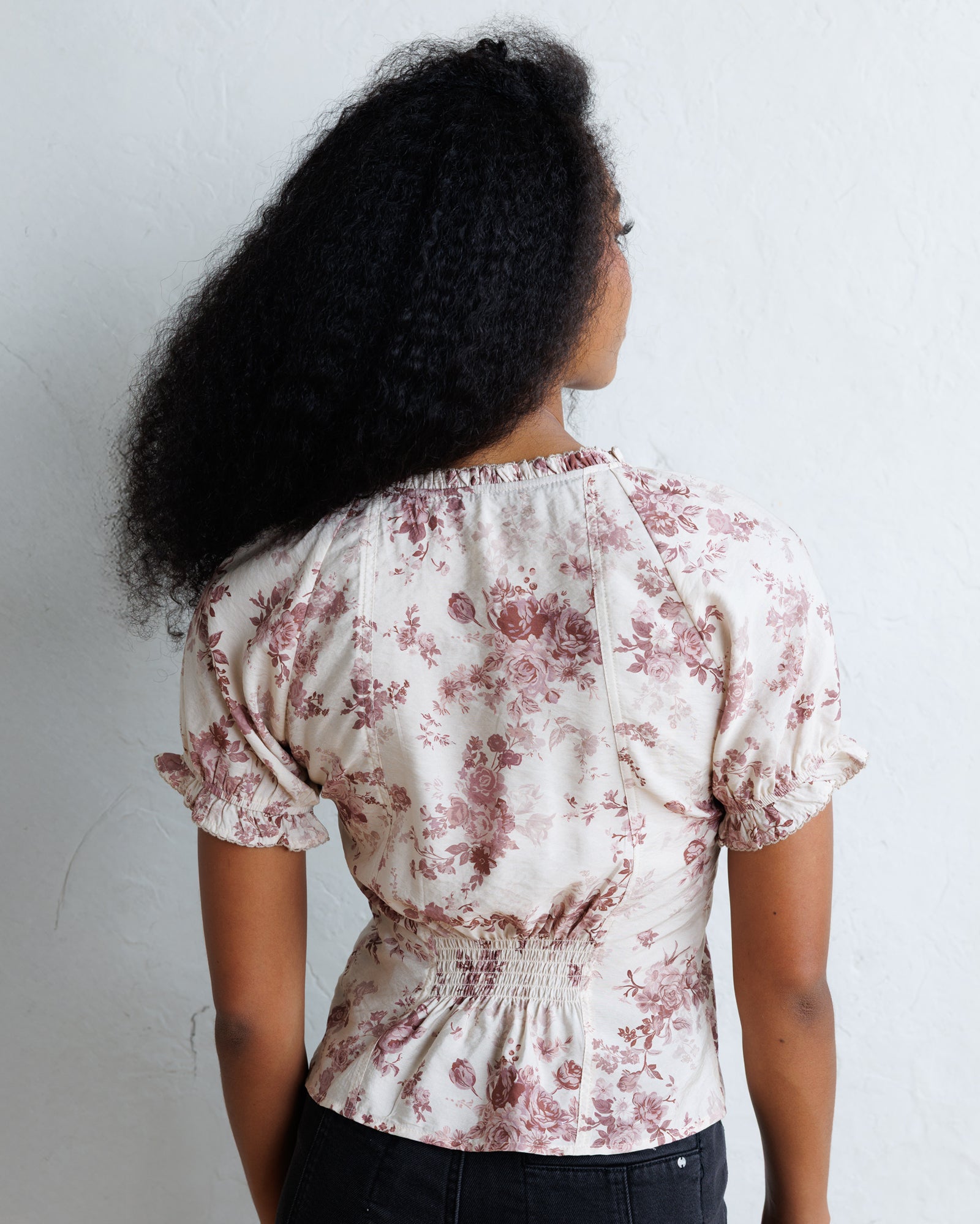 A woman viewed from behind wearing a cream-colored blouse with a dusty rose floral pattern, short puff sleeves, and a smocked waist, paired with black pants against a white wall.