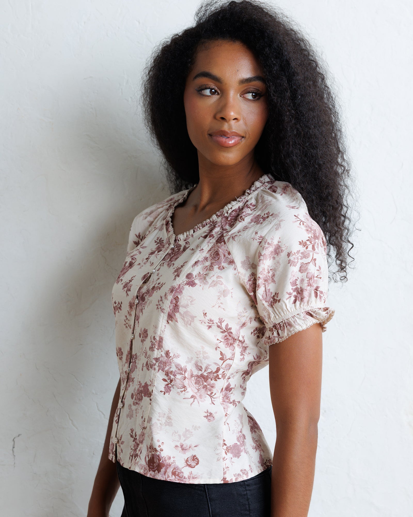 A woman with curly black hair wearing a short-sleeve cream blouse with a dusty rose floral print and puff sleeves, standing against a white textured wall.