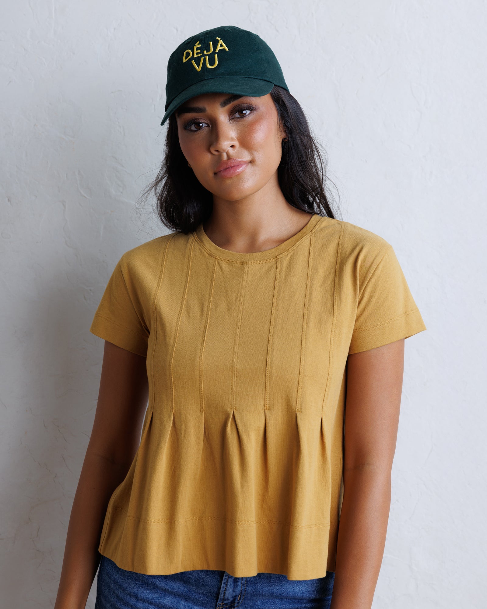 A woman wearing a mustard yellow short-sleeve peplum top, blue jeans, and a dark green baseball cap with yellow text, standing against a textured white wall.
