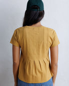 A woman viewed from behind wearing a mustard yellow short-sleeve top with vertical pleats down the back, paired with blue jeans and a dark green baseball cap, against a light gray textured wall.