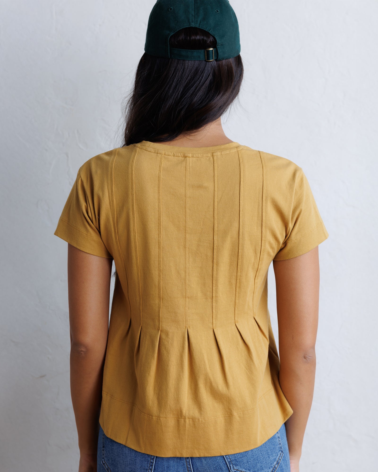 A woman viewed from behind wearing a mustard yellow short-sleeve top with vertical pleats down the back, paired with blue jeans and a dark green baseball cap, against a light gray textured wall.
