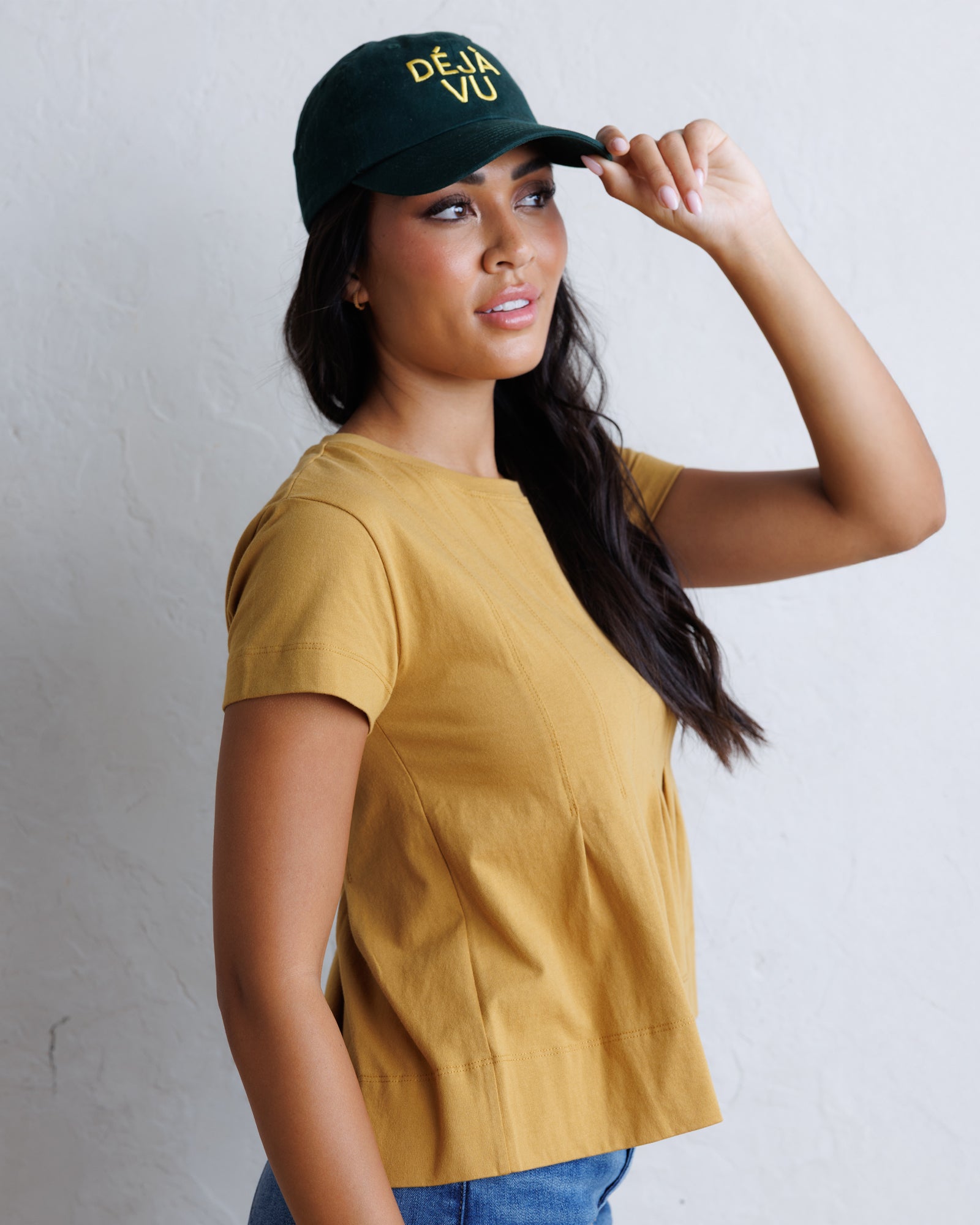A woman wearing a mustard yellow short-sleeve top and a dark green baseball cap with yellow text reading 'ÉDÉN VÙ' poses against a light gray textured wall.