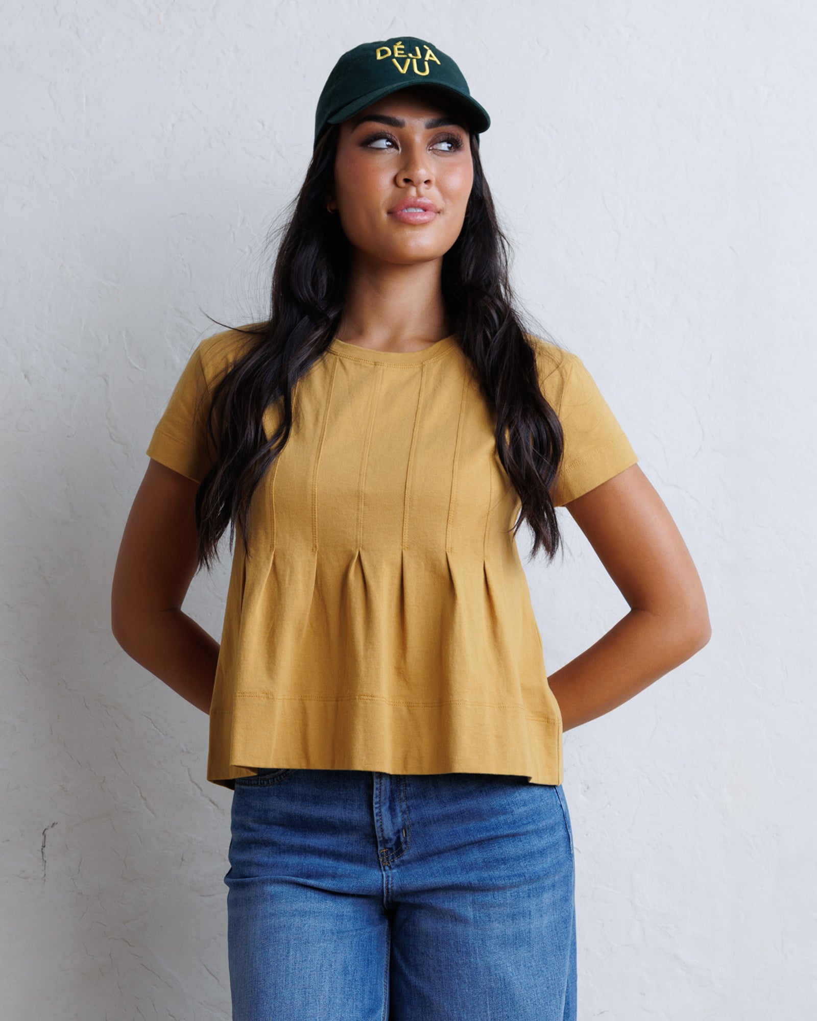 A woman wearing a mustard yellow short-sleeve peplum top, blue jeans, and a dark green baseball cap with yellow text, standing against a textured white wall.