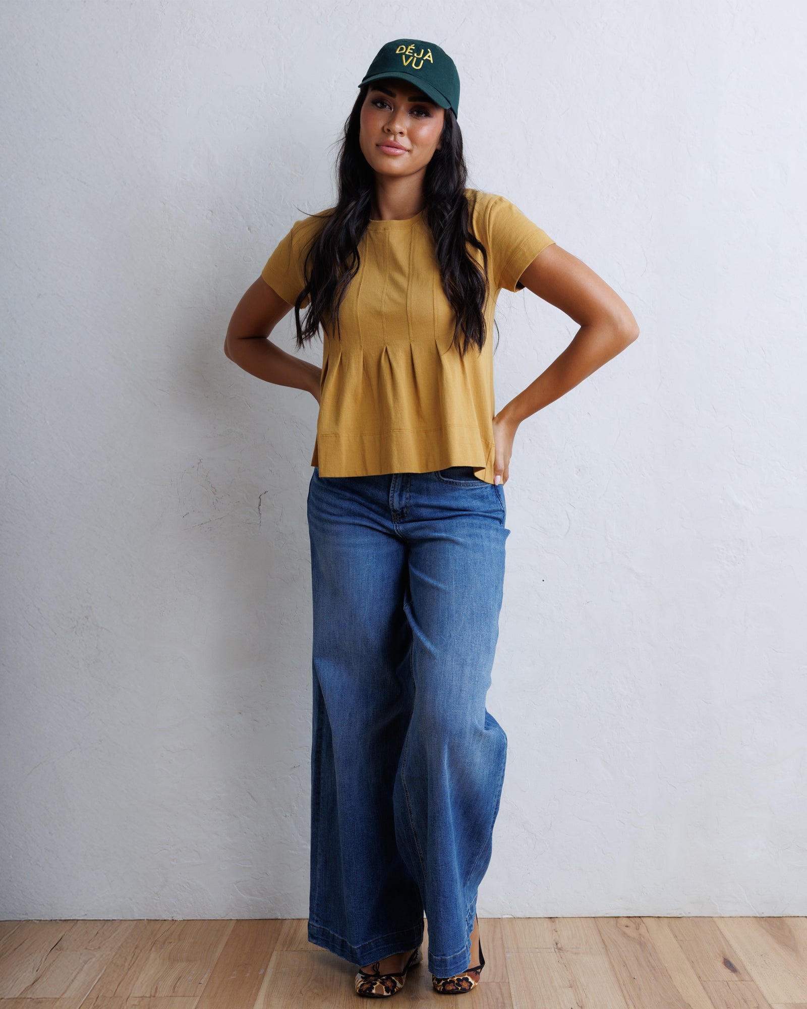 A woman modeling a mustard yellow short-sleeve peplum top, blue wide-leg jeans, and a dark green baseball cap with yellow lettering, standing against a white textured wall on a light wood floor.