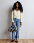 A woman stands against a white wall wearing a pale yellow long-sleeve button-front top, light blue jeans, and black sandals, while holding a patterned gray handbag with black handles.