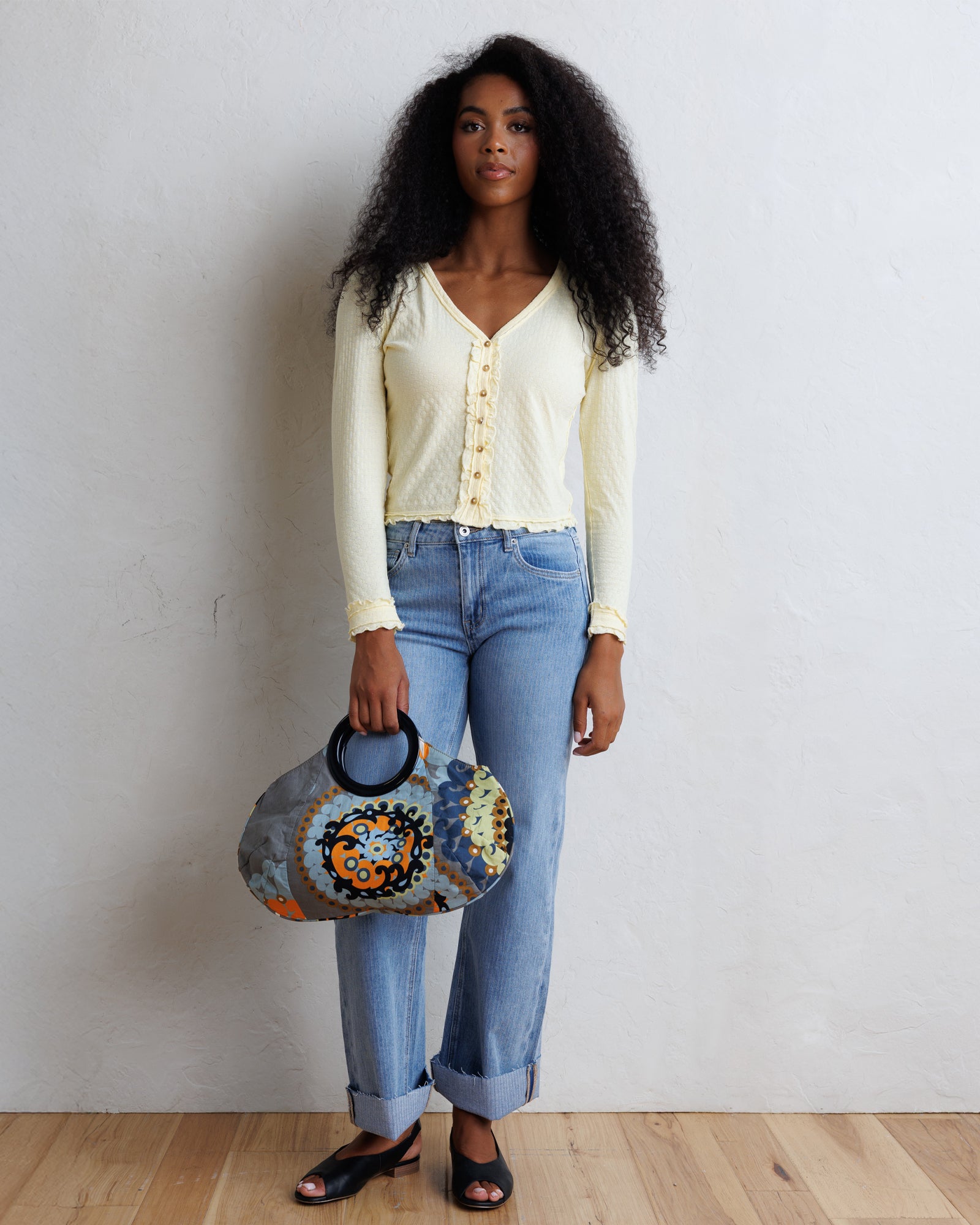 A woman stands against a white wall wearing a pale yellow long-sleeve button-front top, light blue jeans, and black sandals, while holding a patterned gray handbag with black handles.