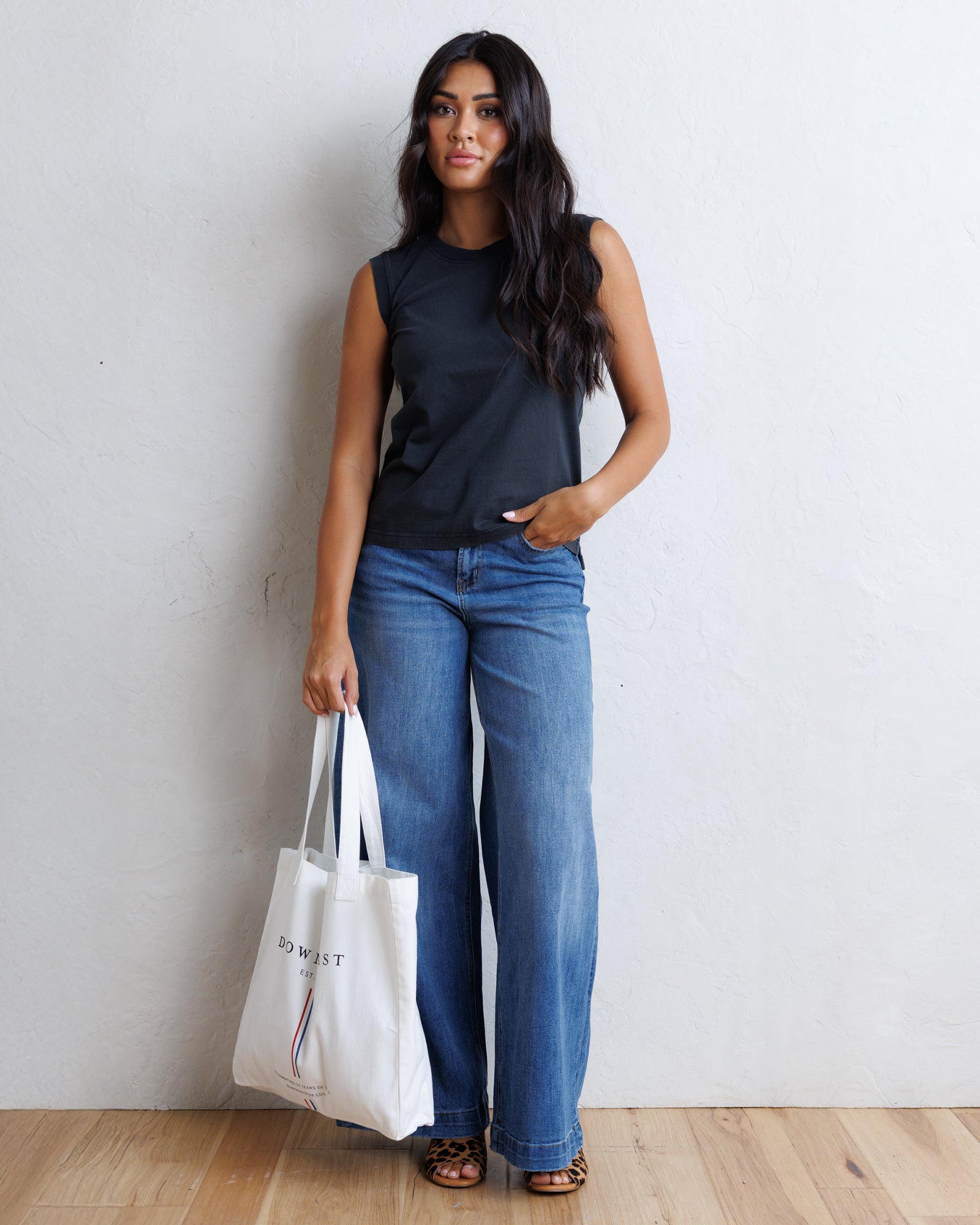A woman stands against a white wall wearing a black sleeveless top, wide-leg blue jeans, and brown sandals, holding a white tote bag with black text.