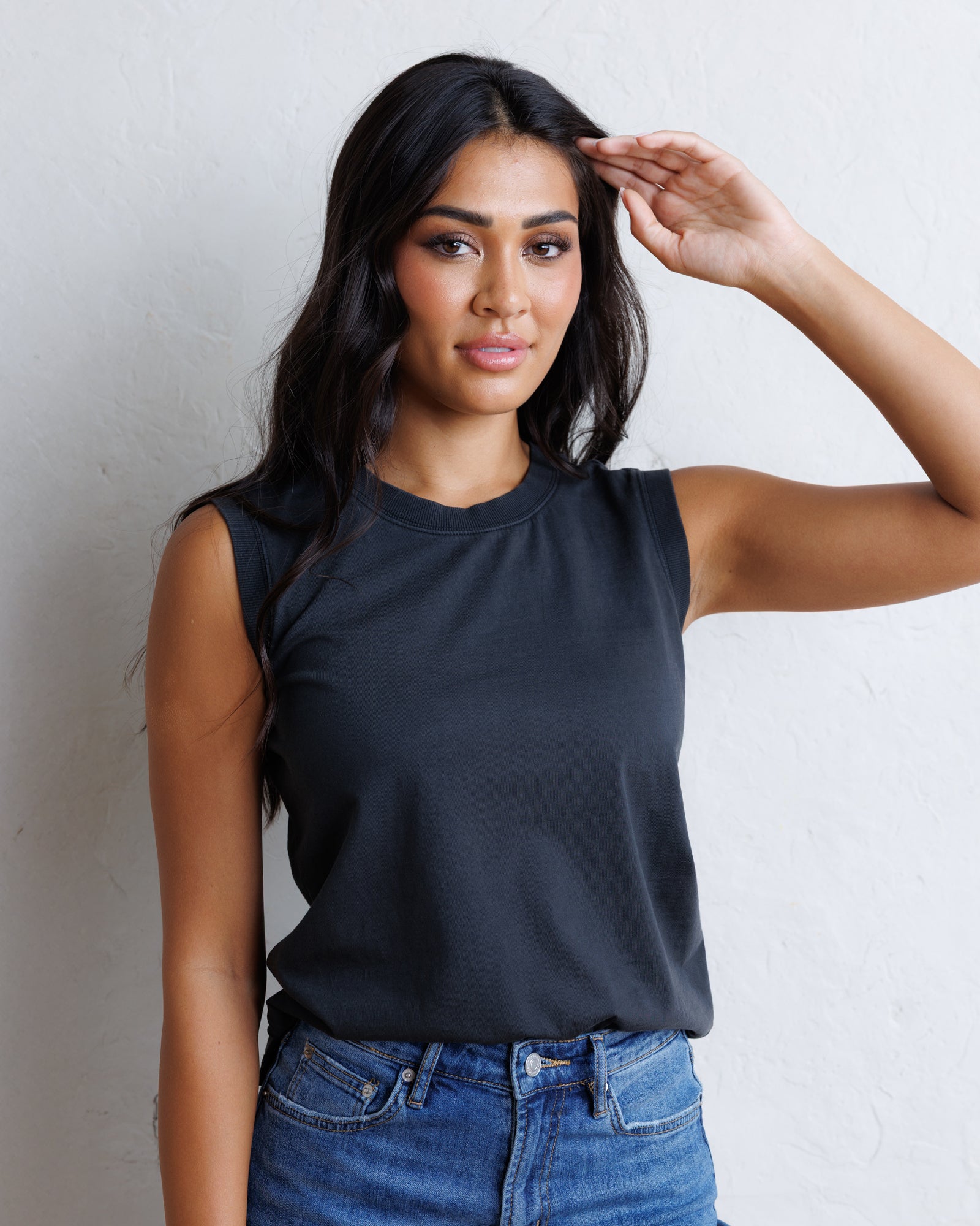 A woman wearing a black sleeveless crewneck t-shirt and blue jeans, posing against a textured white wall.