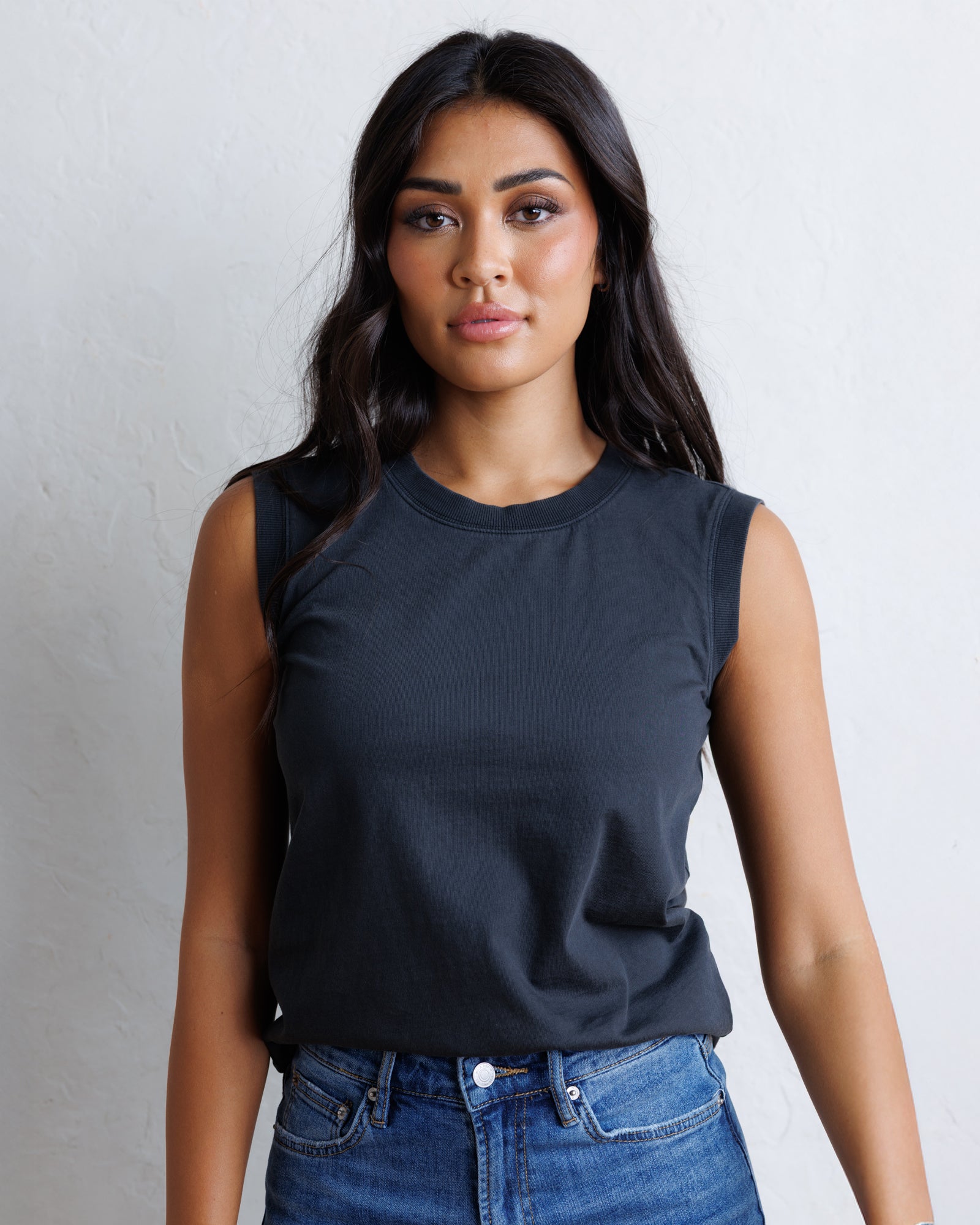 A woman with long black hair wearing a black sleeveless top and blue jeans, standing against a textured off-white wall.