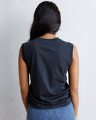 A woman viewed from behind wearing a black sleeveless t-shirt and blue jeans, standing against a light gray textured wall.