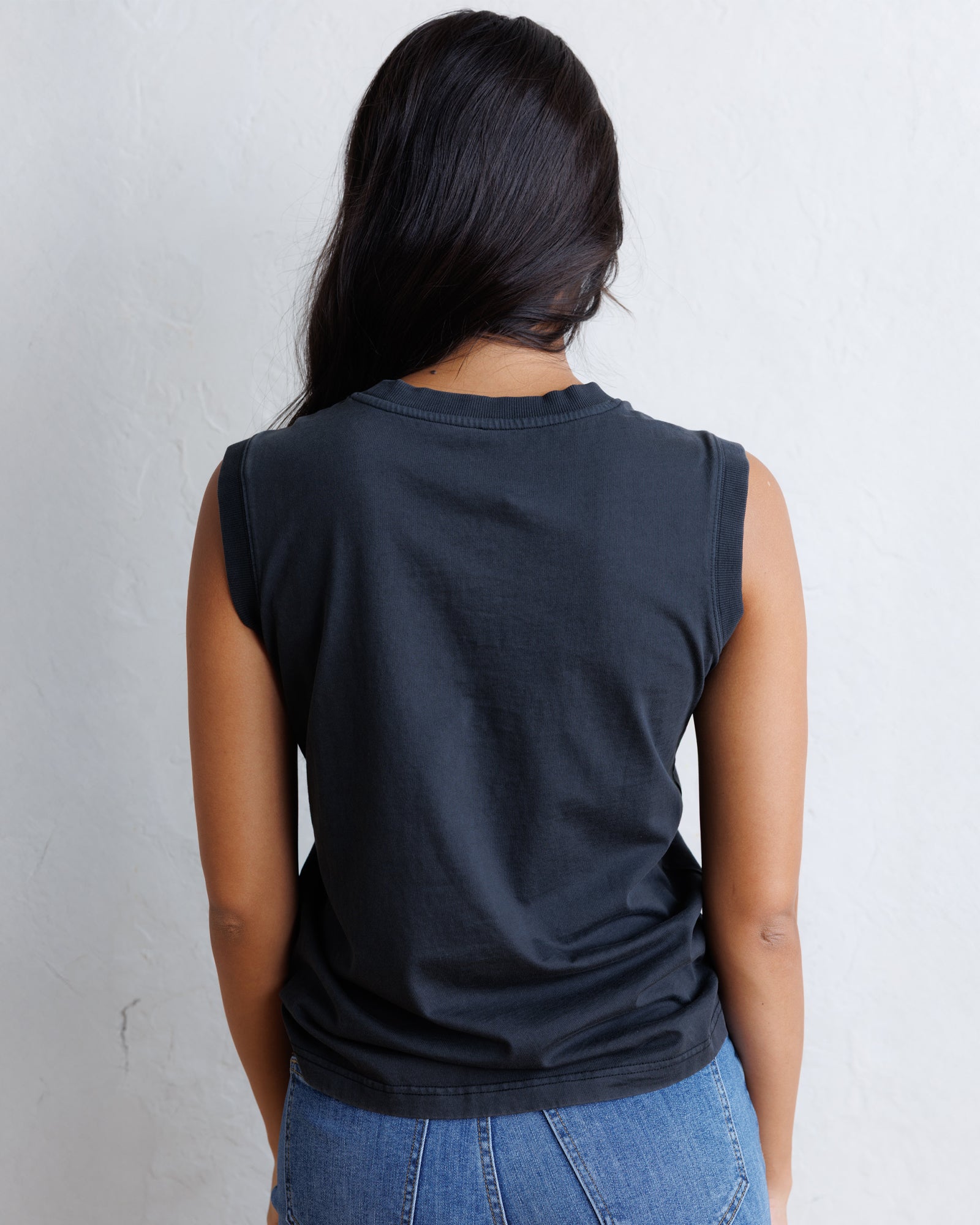 A woman viewed from behind wearing a black sleeveless t-shirt and blue jeans, standing against a light gray textured wall.