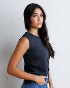 A woman with long black hair wearing a black sleeveless top and blue jeans, standing against a textured off-white wall.