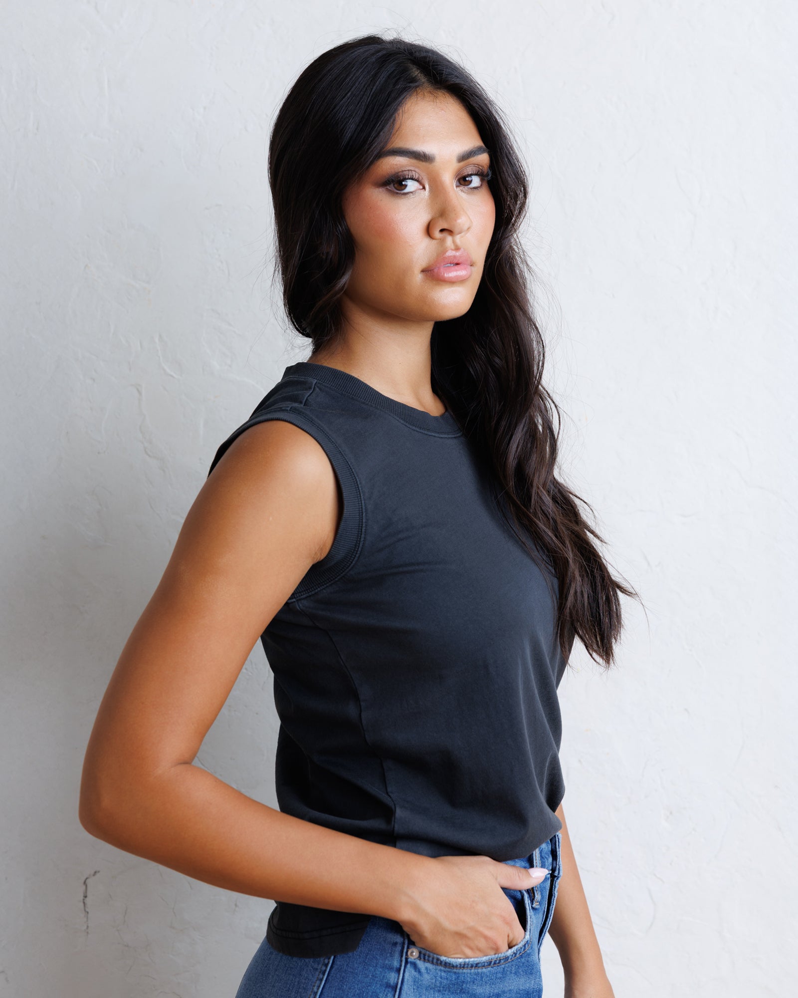 A woman with long black hair wearing a black sleeveless top and blue jeans, standing against a textured off-white wall.