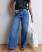A model wearing high-waisted, wide-leg blue denim jeans with a black sleeveless top and leopard-print sandals, holding a white tote bag against a textured white wall and light wood floor.