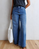 A model wearing high-waisted, wide-leg blue denim jeans with a black sleeveless top and leopard-print sandals, holding a white tote bag against a textured white wall and light wood floor.