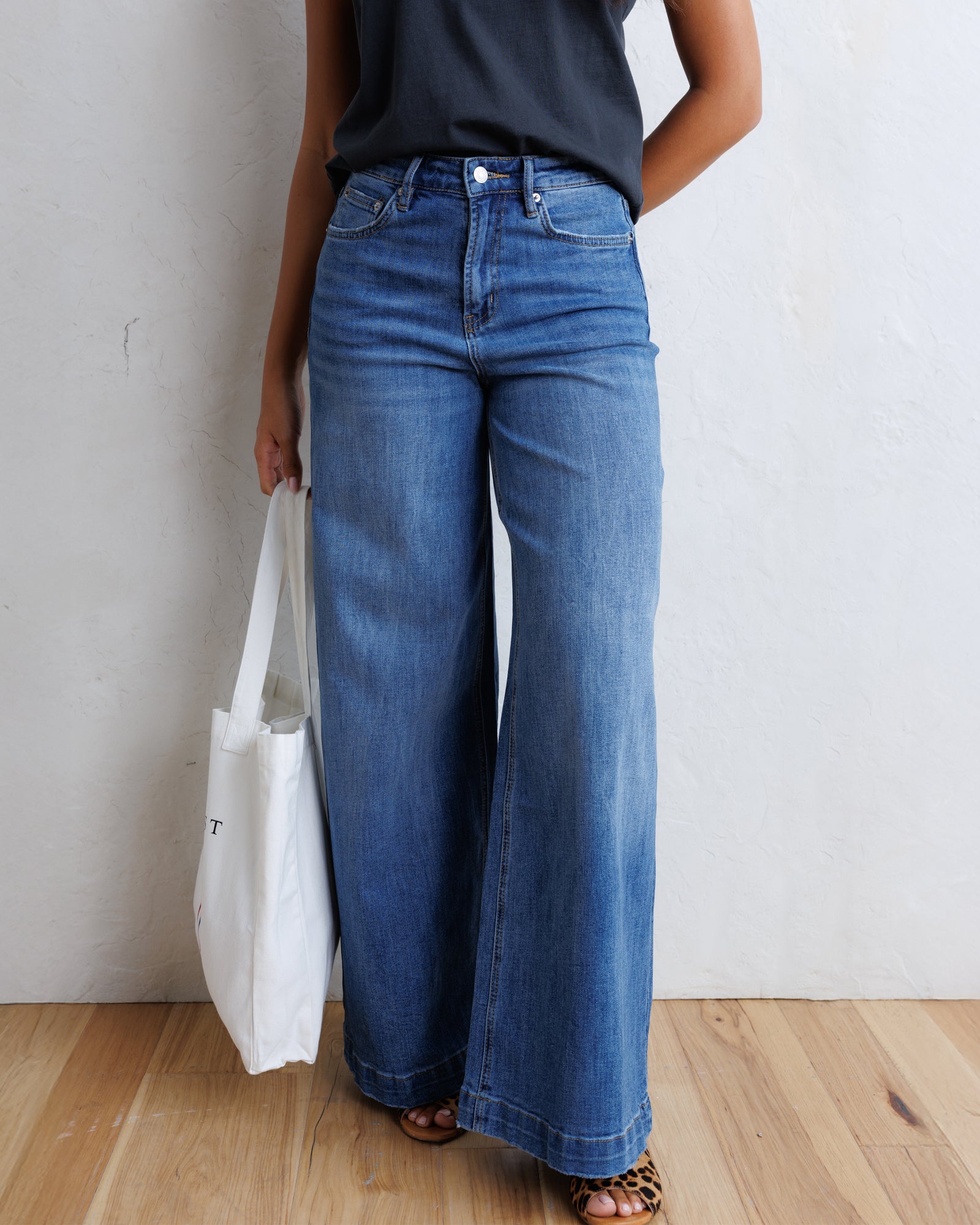 A model wearing high-waisted, wide-leg blue denim jeans with a black sleeveless top and leopard-print sandals, holding a white tote bag against a textured white wall and light wood floor.