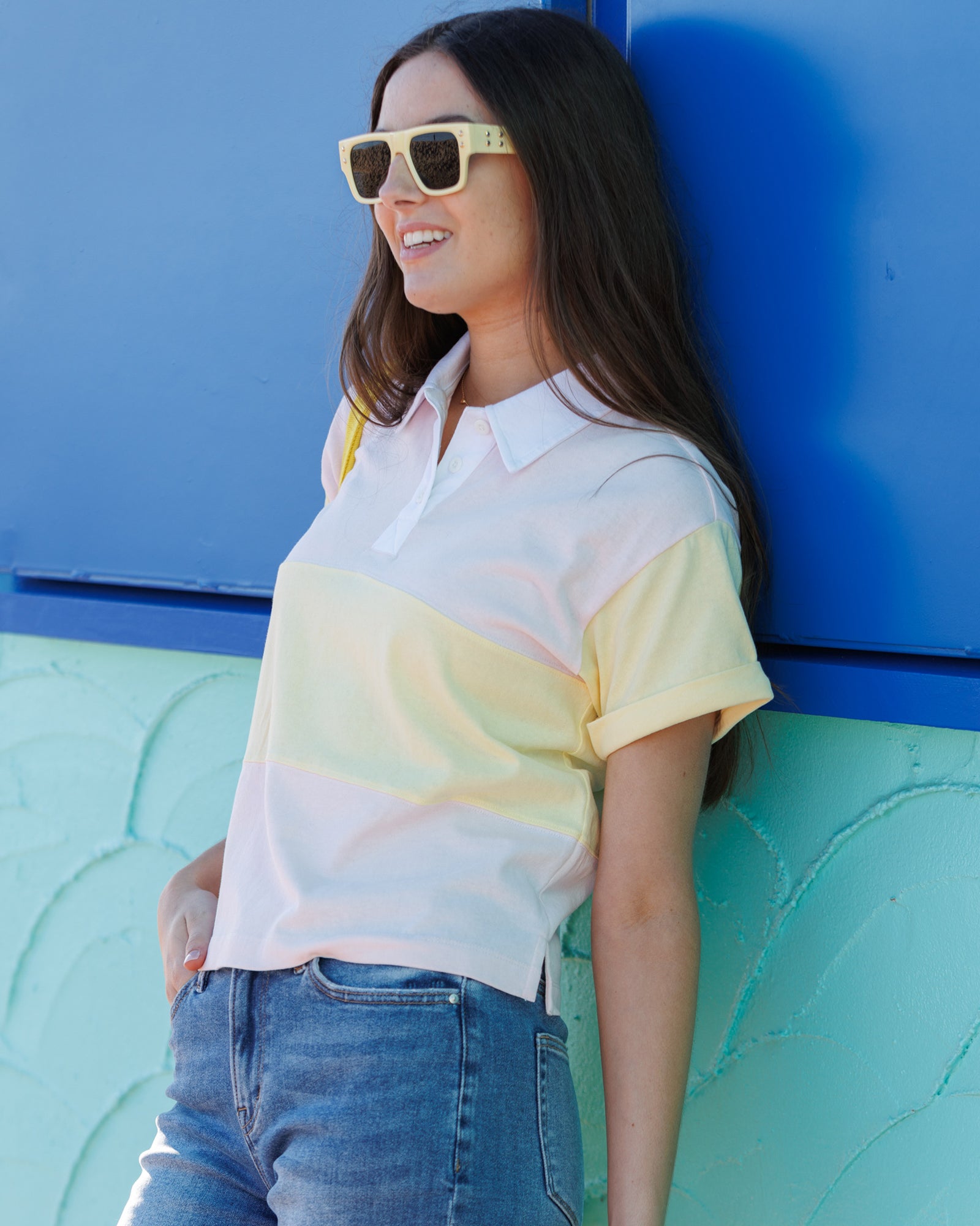 Woman wearing a colorful striped shirt and sunglasses against a blue and green wall.