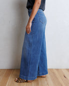 A person wearing a pair of wide-leg blue denim jeans, shown from the side against a textured off-white wall and light wood floor.