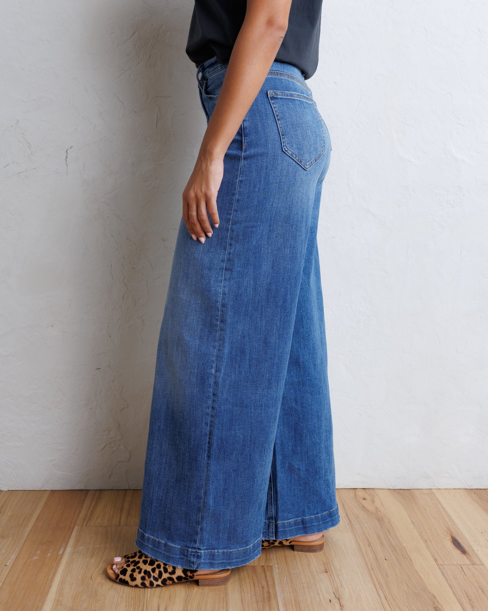 A person wearing a pair of wide-leg blue denim jeans, shown from the side against a textured off-white wall and light wood floor.