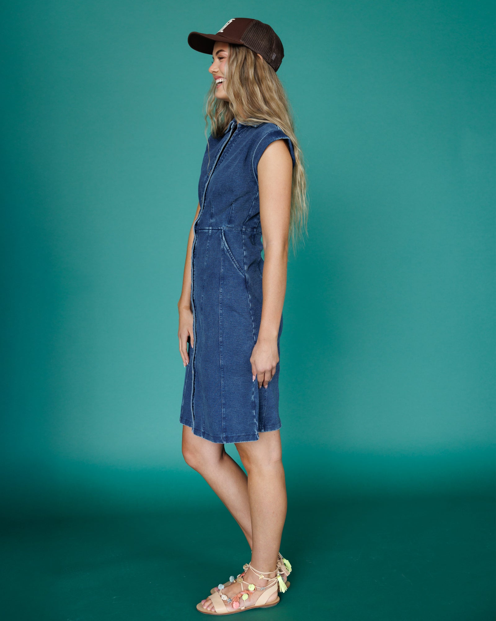 Woman wearing a blue denim dress and brown cap against a teal background