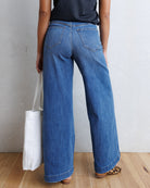 A person shown from the back wearing wide-leg blue denim jeans, a dark top, and leopard-print flat shoes, standing on a wood floor against a light textured wall and holding a white tote bag.