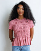 A woman with long, curly black hair wearing a dusty rose pink smocked short-sleeve top and light blue jeans, standing against a white wall.