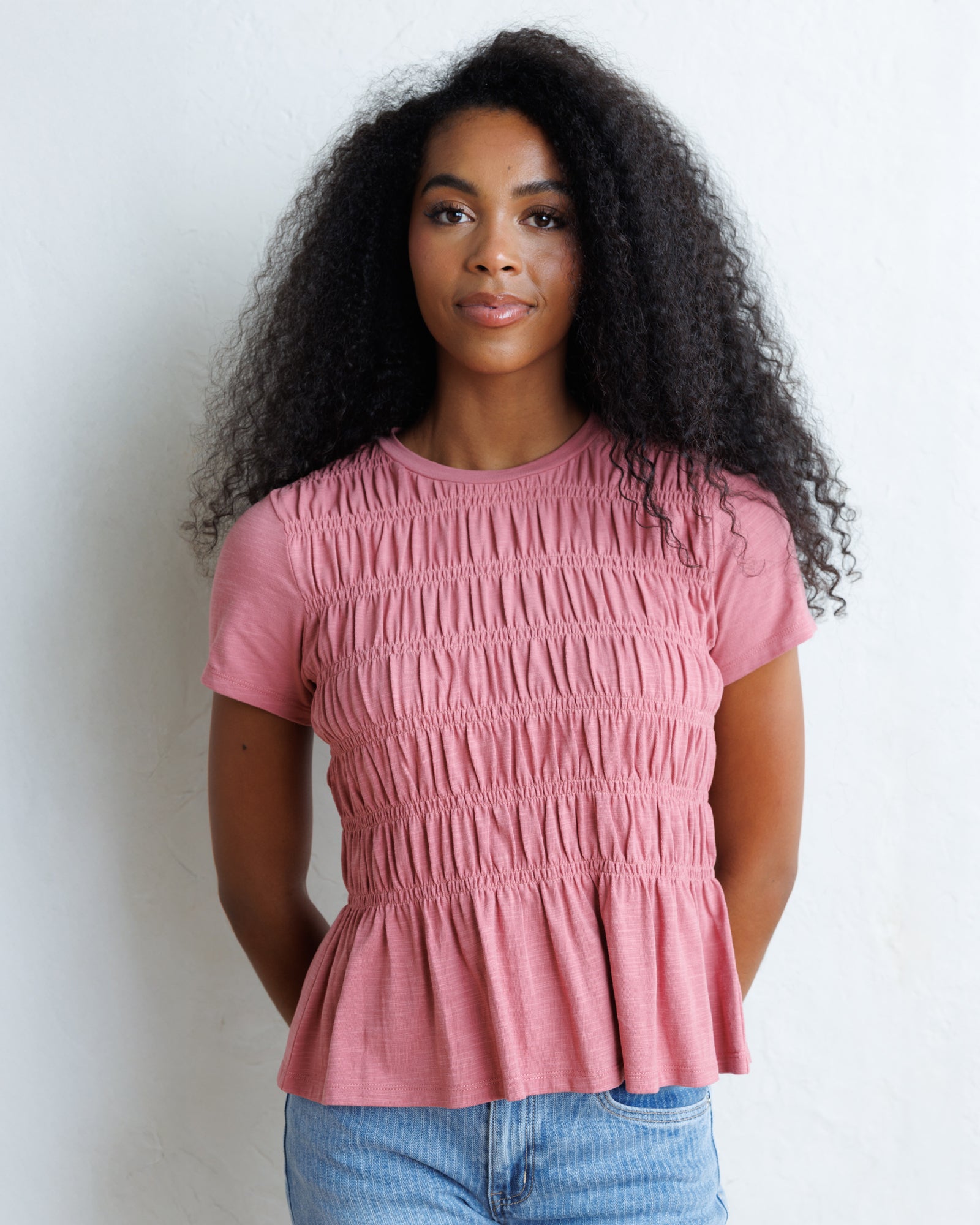 A woman with long, curly black hair wearing a dusty rose pink smocked short-sleeve top and light blue jeans, standing against a white wall.