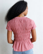 A woman with long, curly black hair wearing a dusty rose pink smocked short-sleeve top and light blue jeans, standing against a white wall.