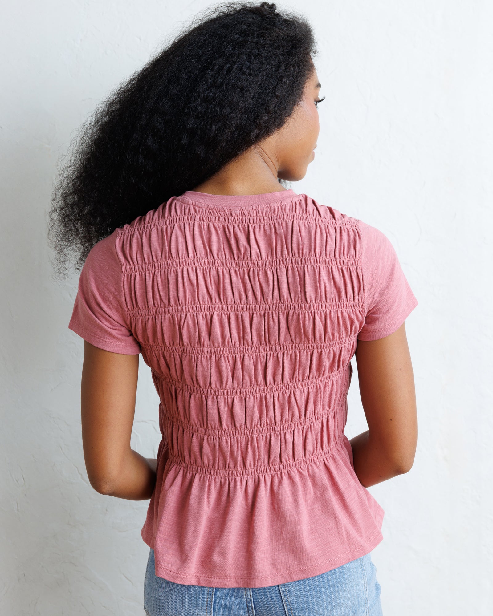A woman with long, curly black hair wearing a dusty rose pink smocked short-sleeve top and light blue jeans, standing against a white wall.