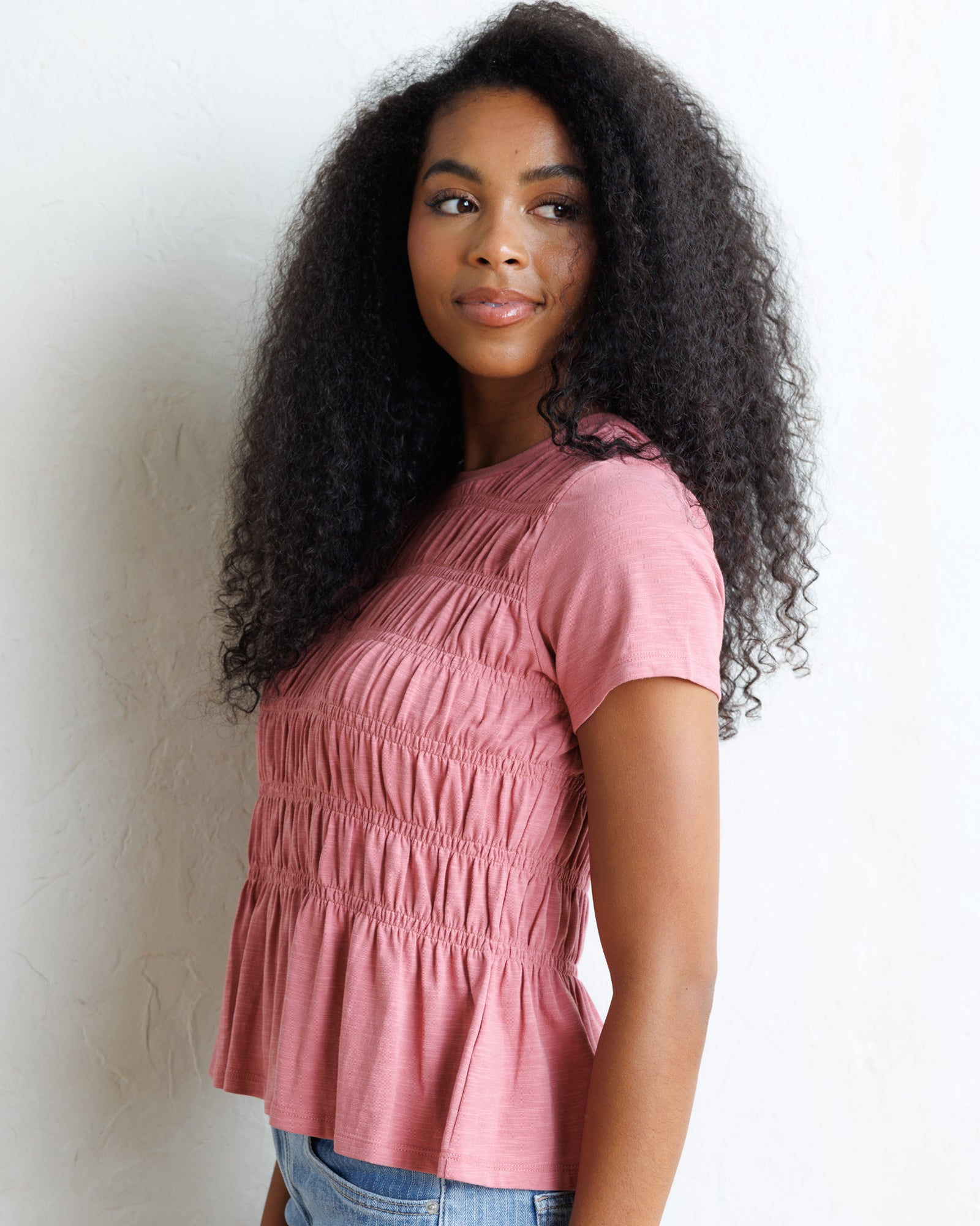 A woman with long, curly black hair wearing a dusty rose pink short-sleeve top with horizontal smocking and a peplum hem, paired with blue jeans, standing against a white textured wall.