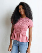 A woman with long, curly black hair wearing a dusty rose pink smocked short-sleeve top and light blue jeans, standing against a white wall.