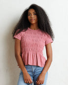 A woman with long, curly black hair wearing a dusty pink short-sleeve smocked peplum top and light blue jeans, standing against a white textured wall.