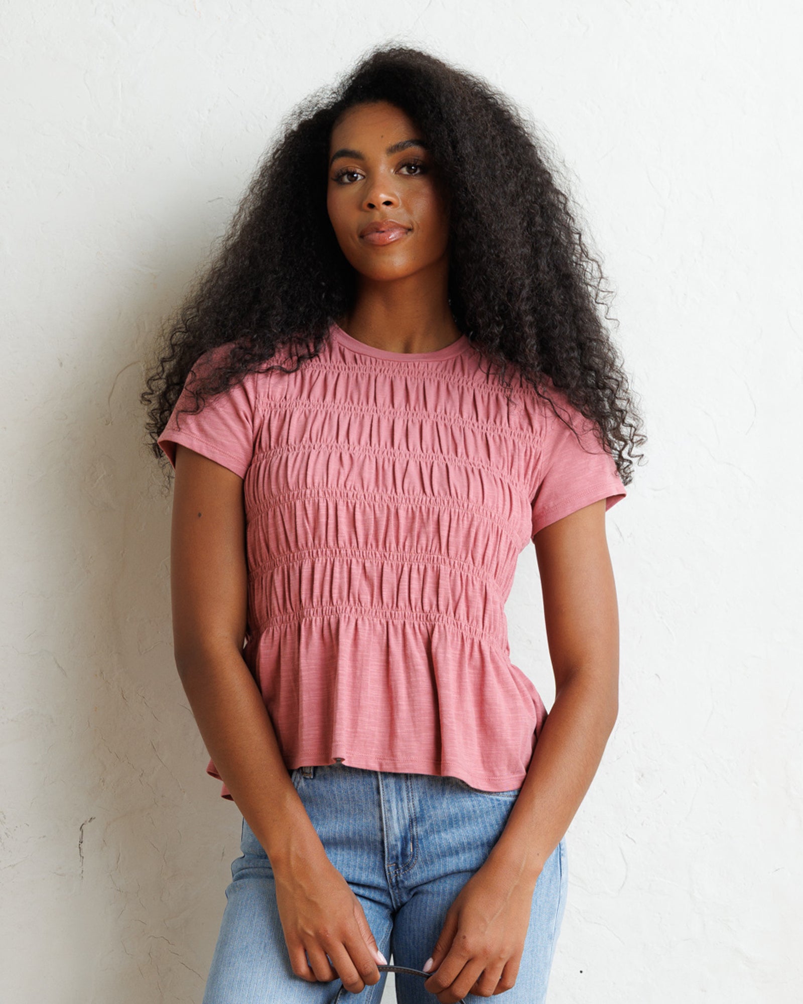A woman with long, curly black hair wearing a dusty pink short-sleeve smocked peplum top and light blue jeans, standing against a white textured wall.