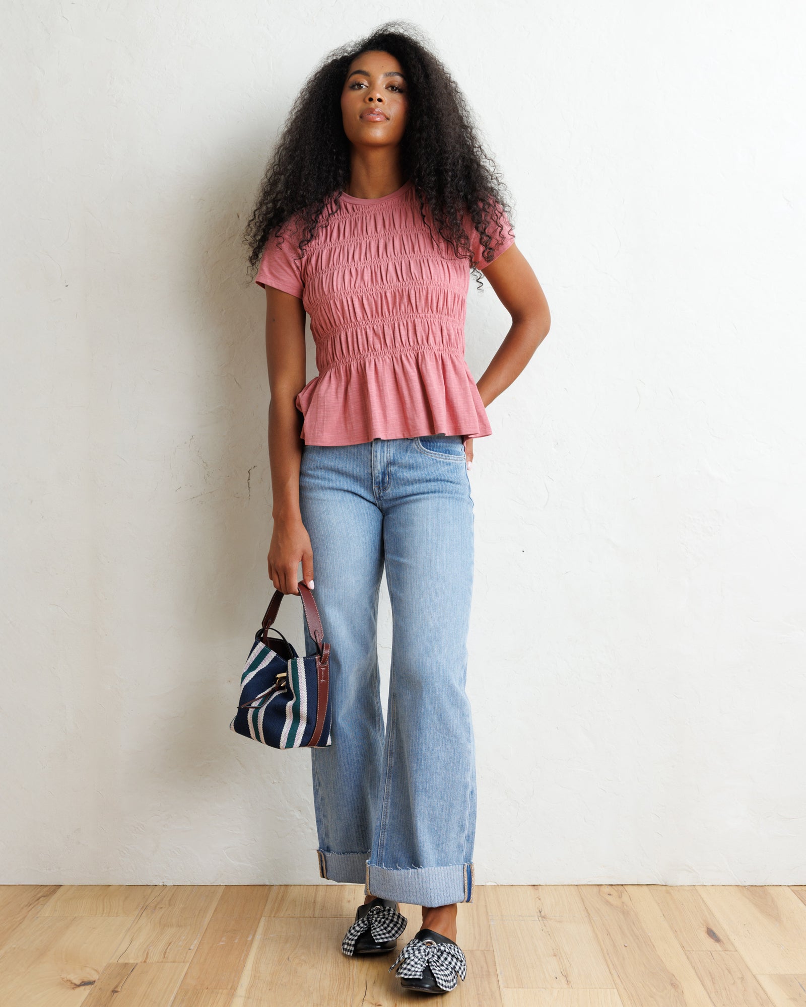 A woman stands against a white wall wearing a pink smocked short-sleeve peplum top, light blue jeans, and patterned loafers, while holding a striped handbag.