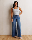 A woman stands against a textured off-white wall on a light wood floor, wearing a light gray ribbed tank top and high-waisted, wide-leg blue jeans with a black studded belt.