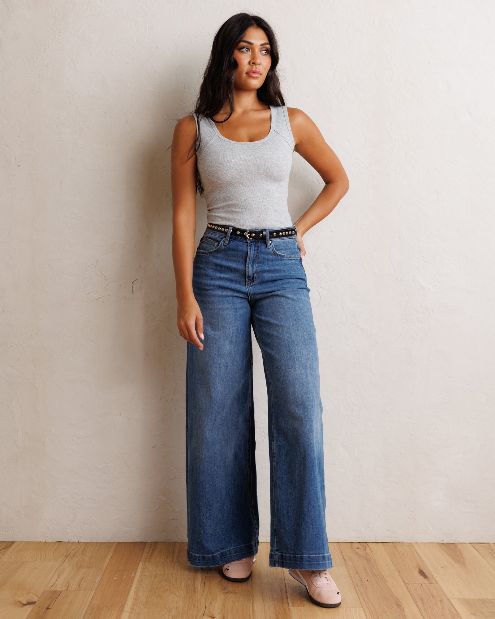 A woman stands against a textured off-white wall on a light wood floor, wearing a light gray ribbed tank top and high-waisted, wide-leg blue jeans with a black studded belt.