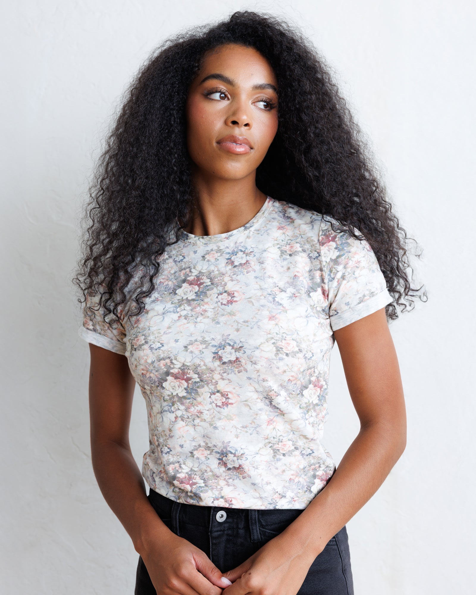 A woman with long, curly black hair wearing a short-sleeve crewneck t-shirt with a delicate pink, blue, and gray floral pattern on a light background, paired with black jeans, set against a plain light gray wall.