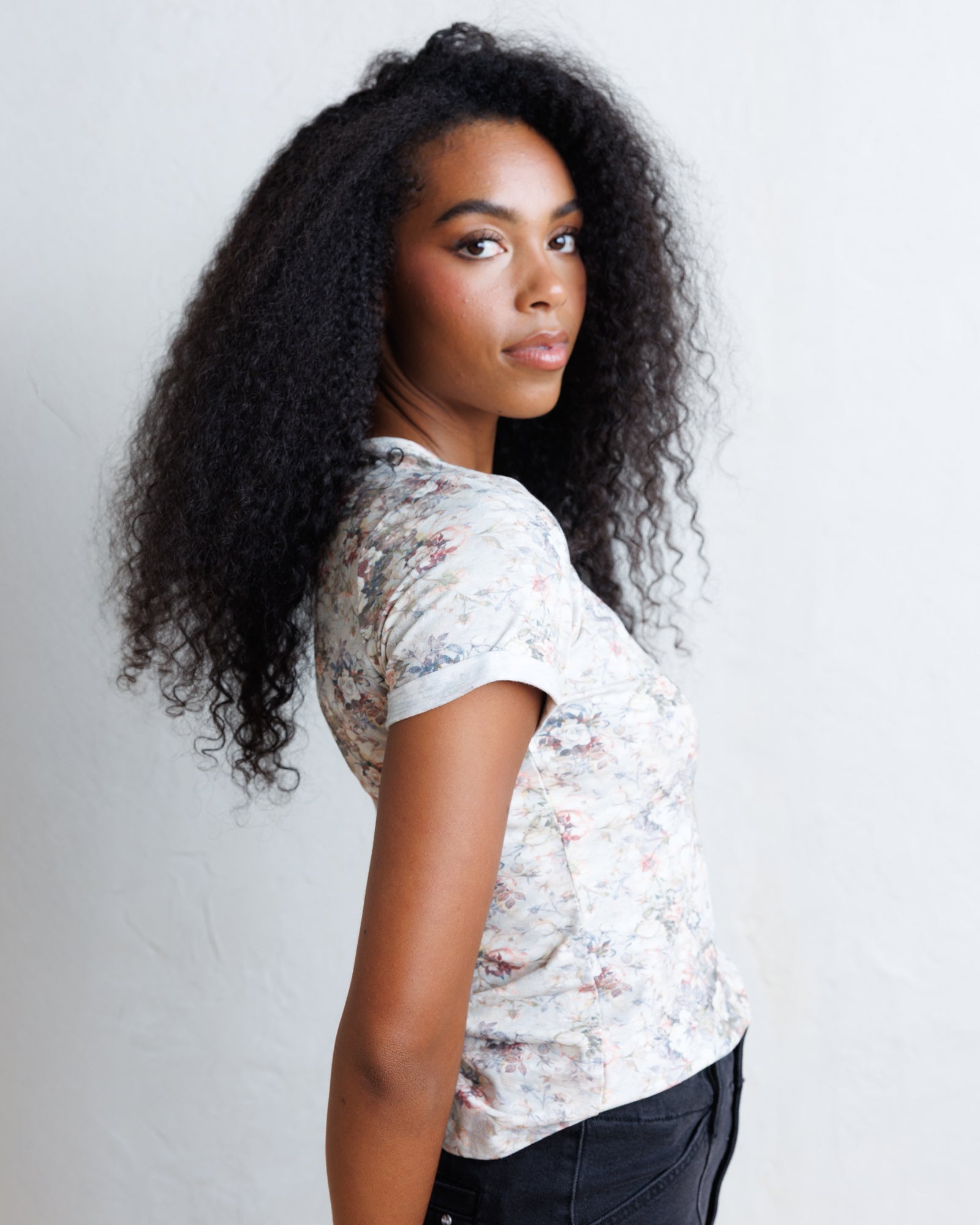 A woman with long, curly black hair is shown from the side, wearing a short-sleeve crewneck t-shirt with a delicate multicolor floral pattern on a light cream background, paired with dark jeans against a plain light gray wall.