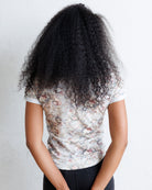 A person shown from behind wearing a short-sleeve t-shirt with a faded floral pattern in muted pink, blue, and gray tones, paired with black pants. The background is a plain white wall.