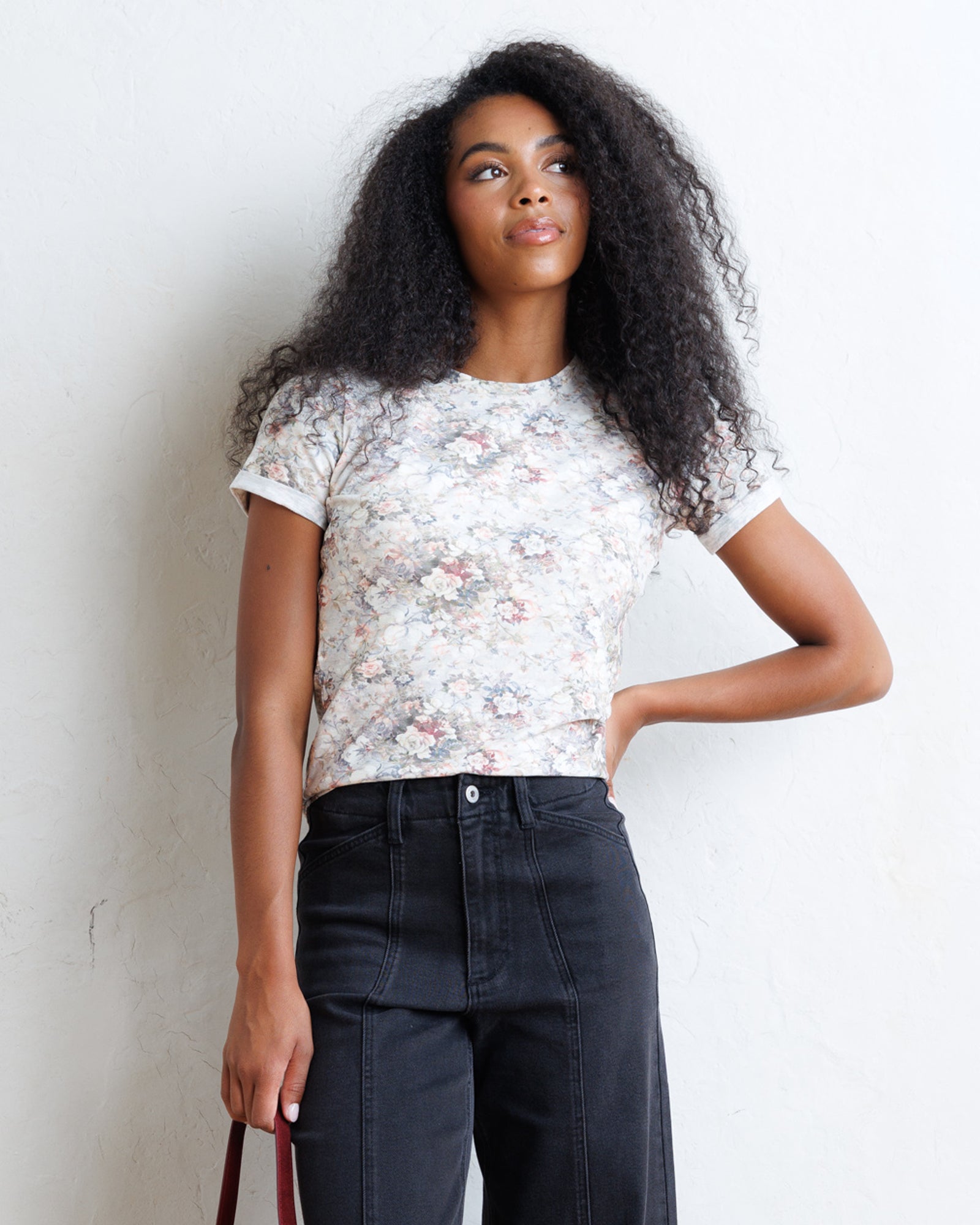 A woman with long, curly black hair models a short-sleeve crewneck t-shirt featuring a delicate white, pink, and green floral pattern, paired with dark charcoal gray jeans, standing against a textured off-white wall.