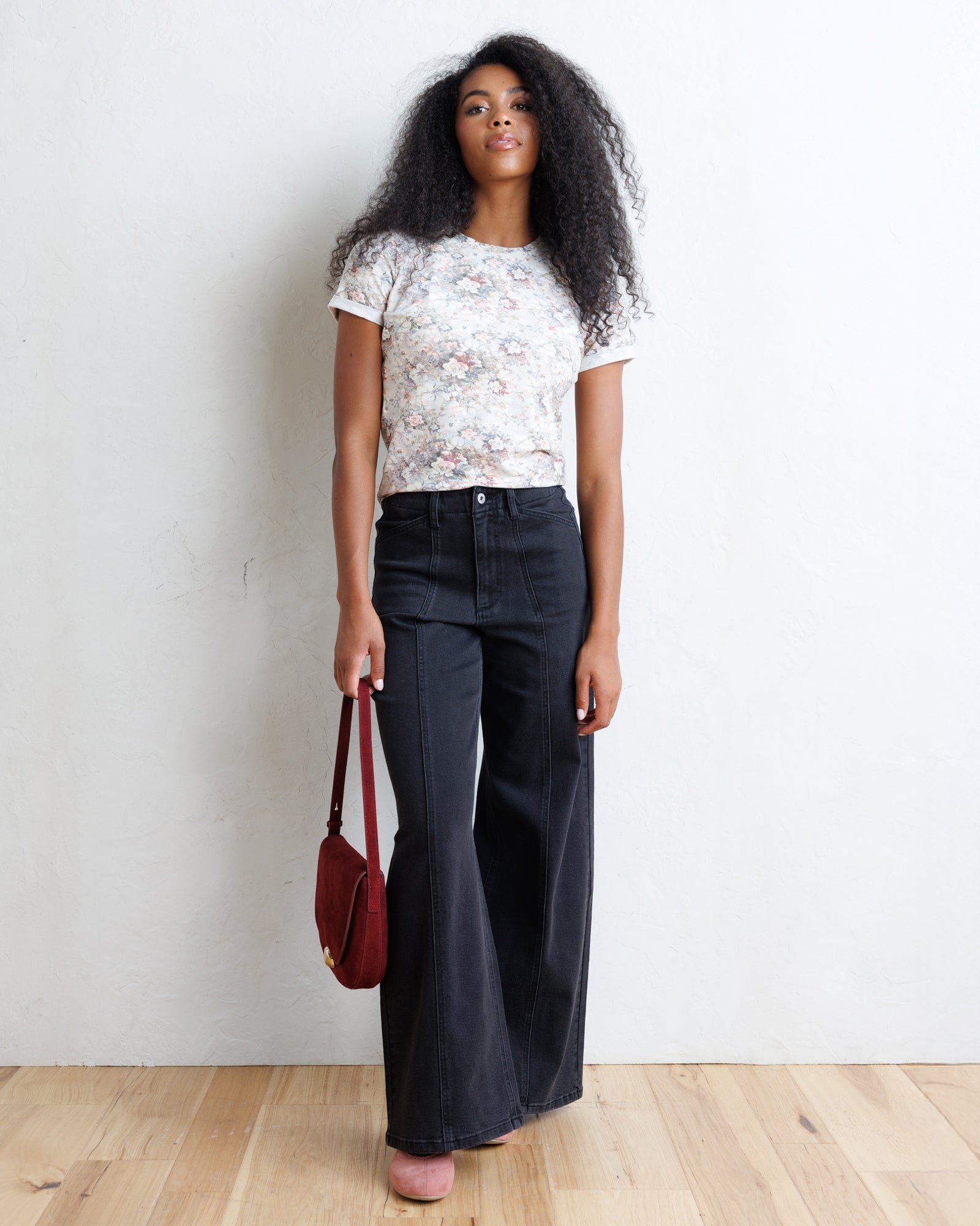 A woman stands against a white wall on a light wood floor, wearing a white short-sleeve floral t-shirt, dark wide-leg jeans, and holding a burgundy shoulder bag.