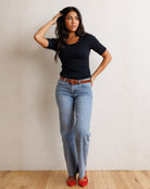 A woman modeling a black short-sleeve t-shirt with a scalloped neckline, paired with light-wash blue jeans, a brown leather belt, and red pointed-toe flats, standing against a textured off-white wall on a light wood floor.