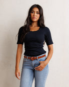 A woman modeling a short-sleeve black top with a scalloped neckline, paired with light blue jeans and a brown belt, standing against a textured off-white wall.
